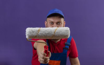 young bearded builder man in construction uniform and cap stretching out to camera paint roller standing over purple background